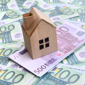 Small toy house is lies on a set of green monetary denominations of 100 euros